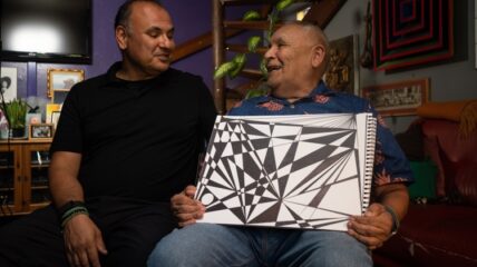 A grown son sits next to his father. The father is holding a drawing he has done.