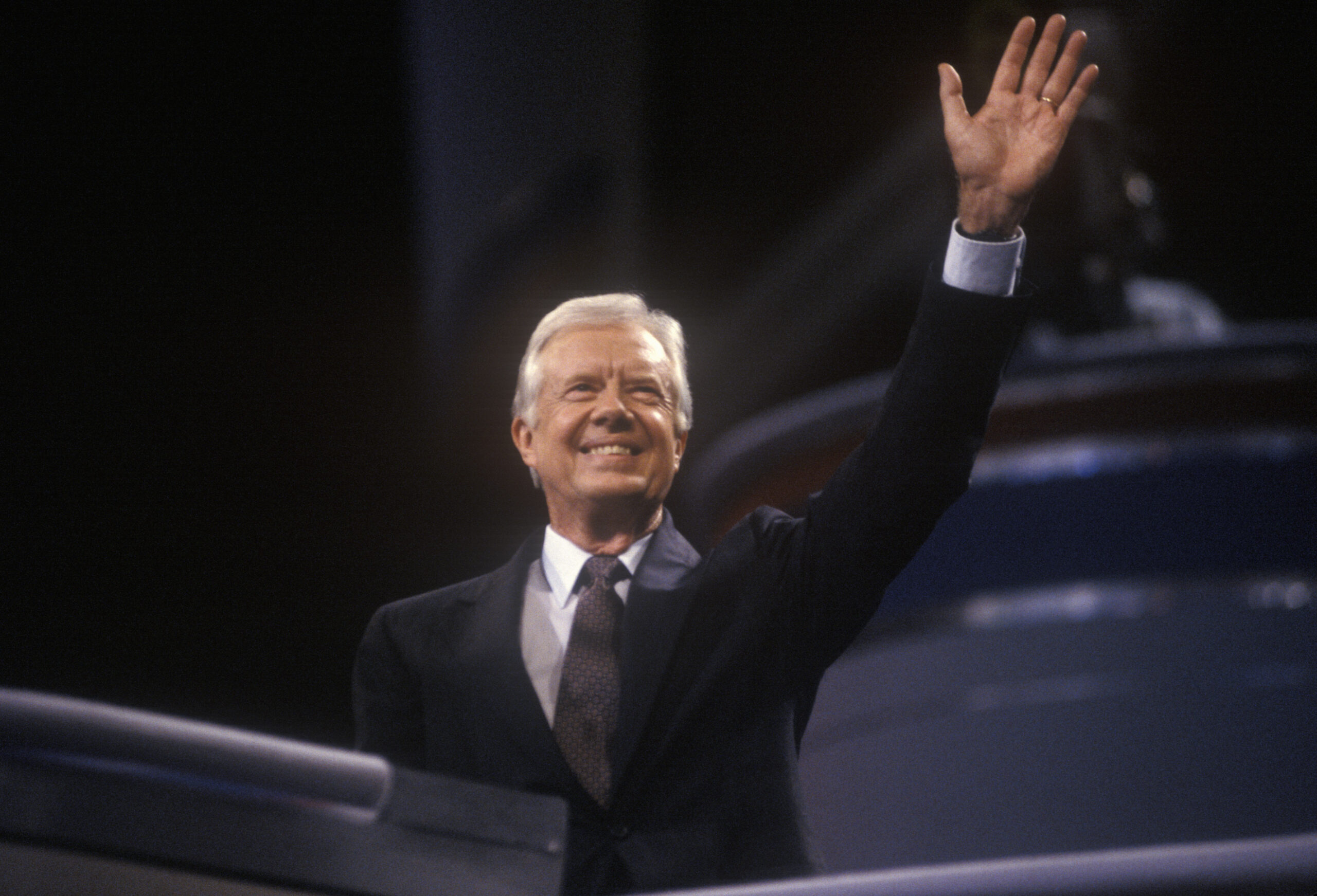 Pres Jimmy Carter in a dark suite, white shirt and tie waving.