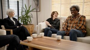 Three people, a white woman with short gray hair in a Black suit, a white woman with short brown hair wearing a Black blouse, and a Black man wearing a flannel shirt all sit in a room together laughing.