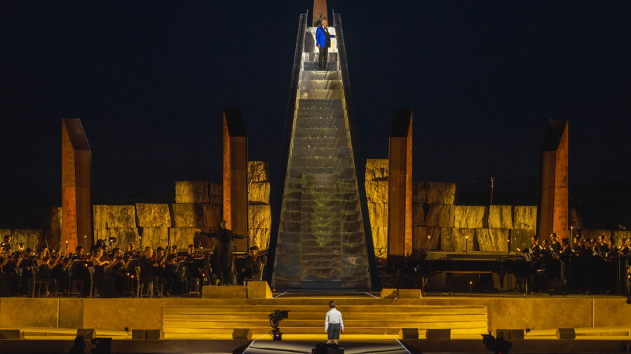 Andrea Bocelli wearing a bright blue blazer and Black pants stands on top of a huge staircase on a stage.