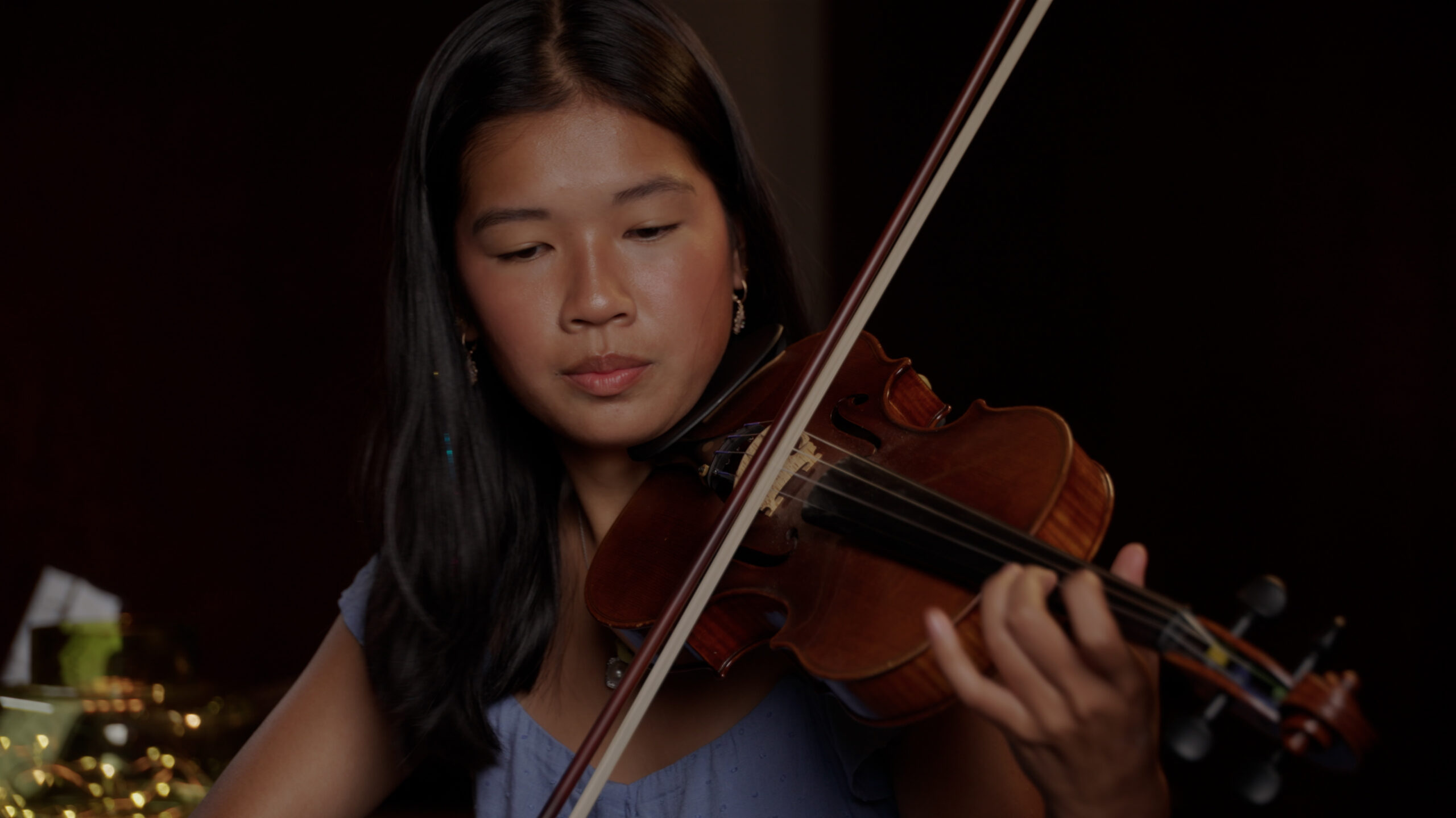 A young woman with long Black hair wearing a purple dress plays the violin.