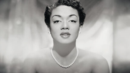 A Black and white photo of Hazel Scott – A Black woman with Black hair pulled off her face. She's wearing pearl earrings and a pearl necklace.