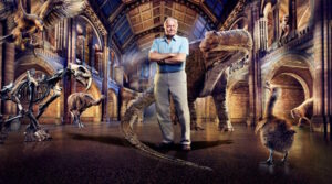 David Attenborough stands with arms folded with dinosaurs all around him