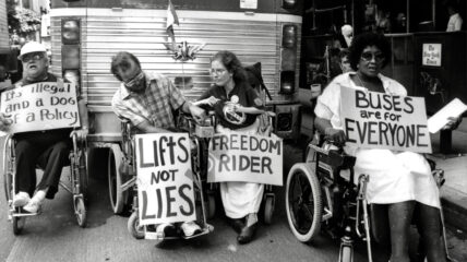 Four people in wheel chairs in front of a bus holding pickets signs that say :Buses are for Everyone," "Lift not lies," and Freedom rider."