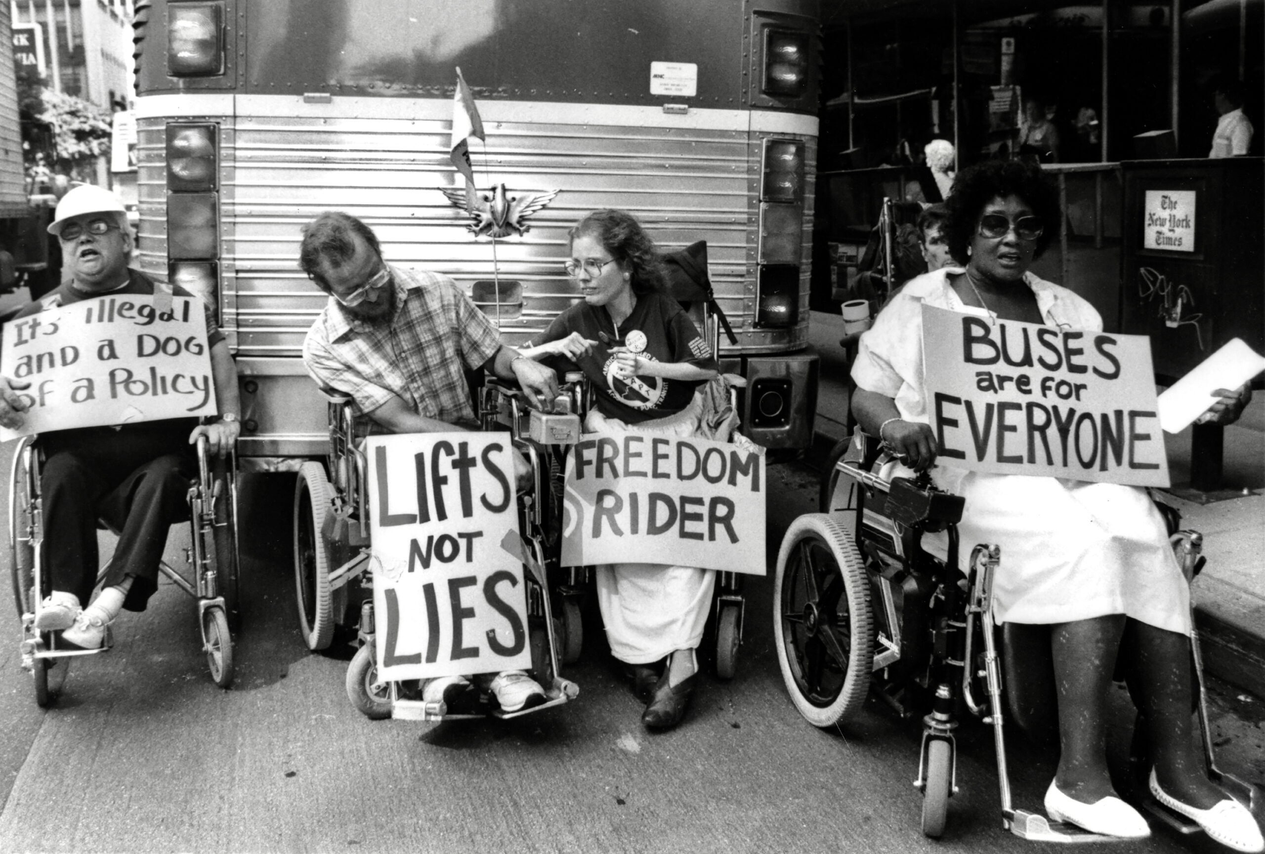 Four people in wheel chairs in front of a bus holding pickets signs that say :Buses are for Everyone," "Lift not lies," and Freedom rider."