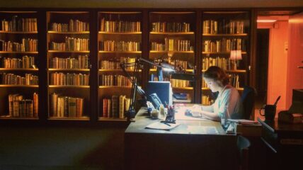 a librarian sits at a desk with a wall of books behind her.