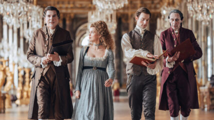 Three men and a woman walk down the hall of a castle with chandeliers lining the ceiling.