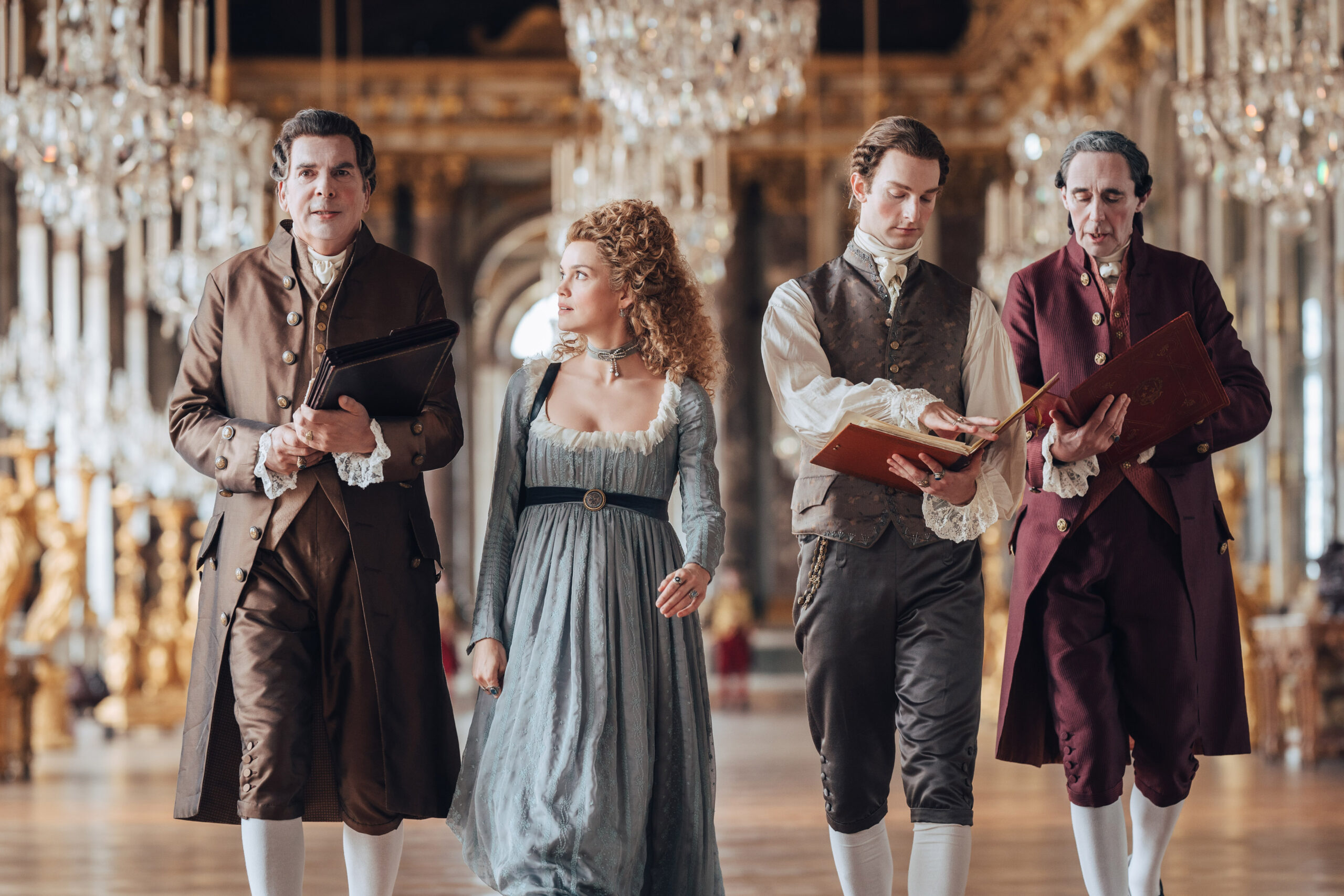 Three men and a woman walk down the hall of a castle with chandeliers lining the ceiling.